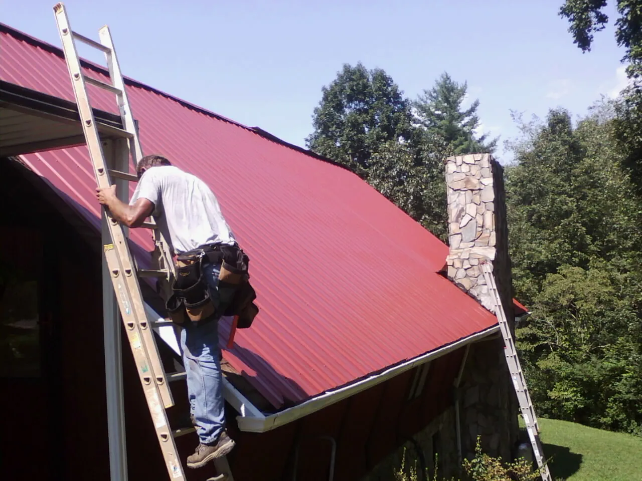Roofer on a ladder beside a red metal roof and stone chimney — Soffit & Fascia Repair by Roofing Brothers in North Tonawanda